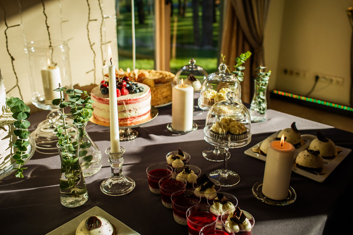 Storos baltos žvakės ir stiklinėmis žvakidėmis dekoruotas saldus stalas